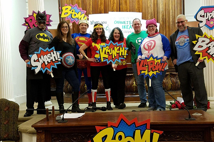 Photo of a group of people wearing masks, superhero shirts and holding up different superhero signs that says "smash," "boom," "krunch," and "kapow." 